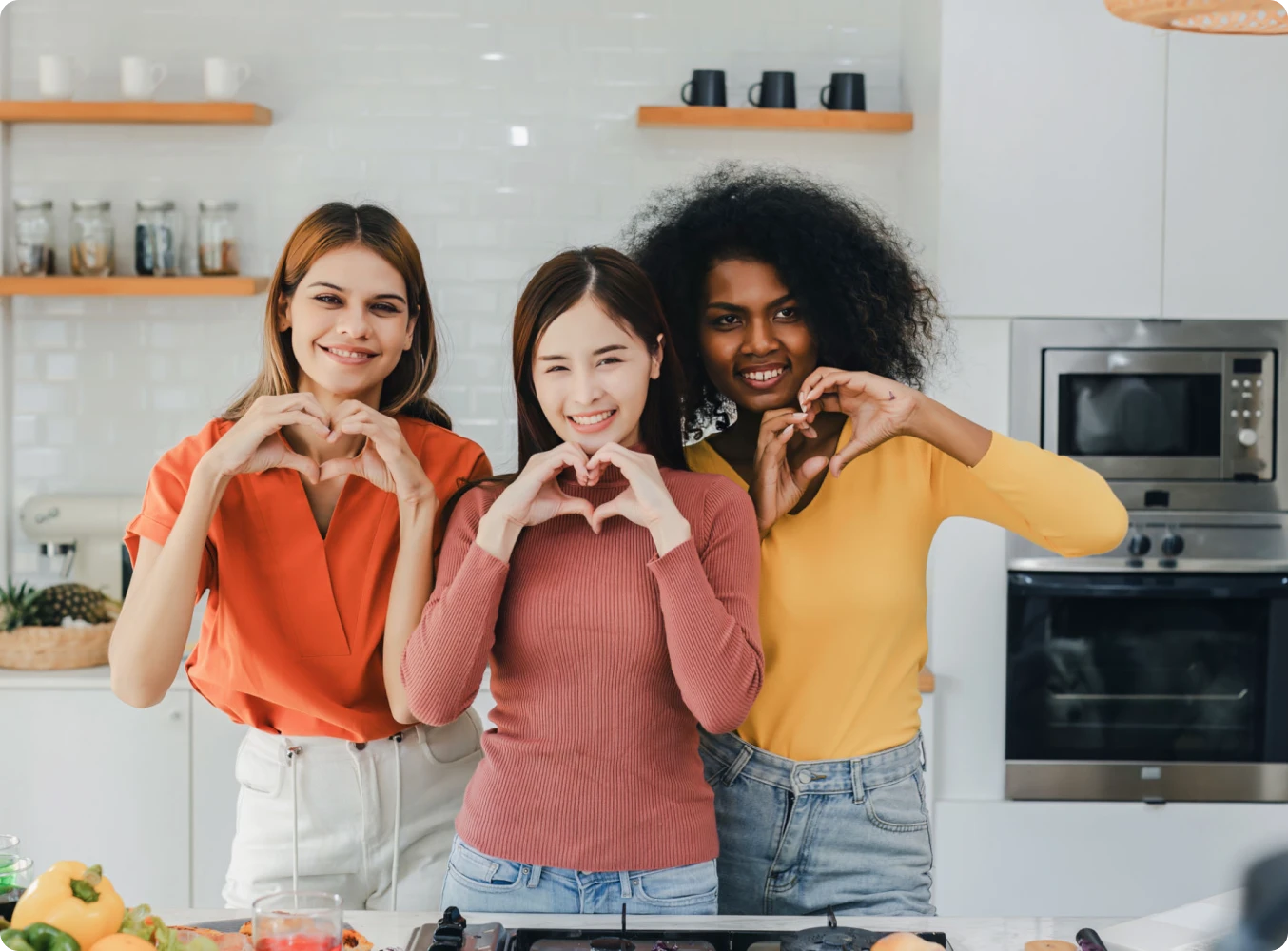 Three women making heart shapes in kitchen.